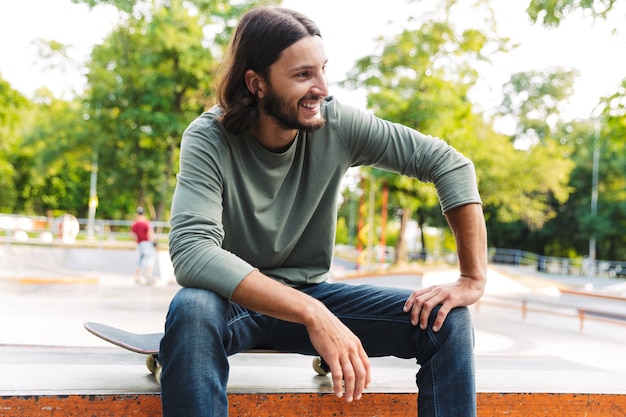 Jeune homme séduisant de hipster assis au skate park avec une planche à roulettes