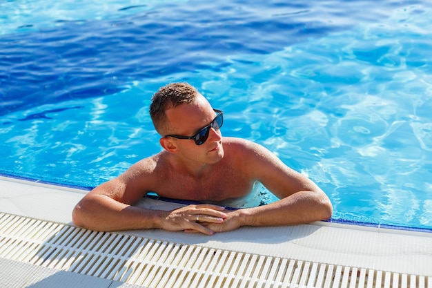 Jeune homme séduisant à lunettes de soleil se repose dans la piscine pendant les vacances d'été. Le gars dans la piscine près de l'hôtel