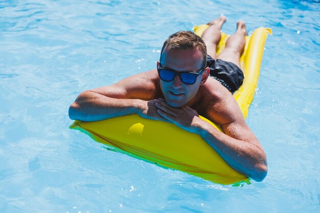 Un jeune homme séduisant en lunettes de soleil et en short se détend sur un matelas jaune gonflable dans la piscine Vacances d'été