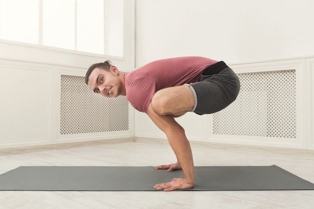 Jeune homme séduisant pratiquant le yoga, debout à Bakasana, faisant des exercices d'équilibre sur un tapis en cours de fitness, vue latérale, espace de copie