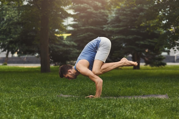 Jeune homme séduisant pratiquant le yoga, debout dans l'exercice Bakasana sur l'herbe verte dans le parc, vue latérale, espace pour copie