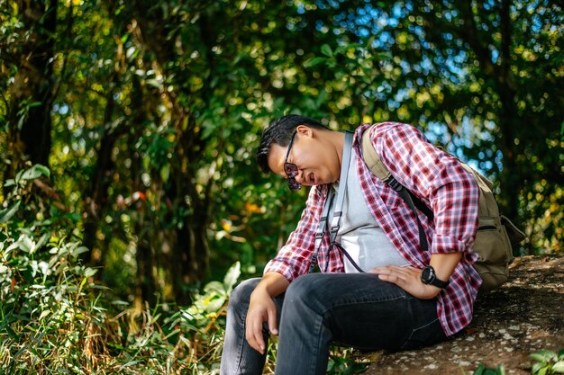 Jeune homme séduisant avec sac à dos de voyage se sentant essayé après une randonnée dans la nature et un environnement frais lors d'un voyage de trekking assis pour se reposer sur des rochers et essuyer la sueur dans l'espace de copie de la forêt