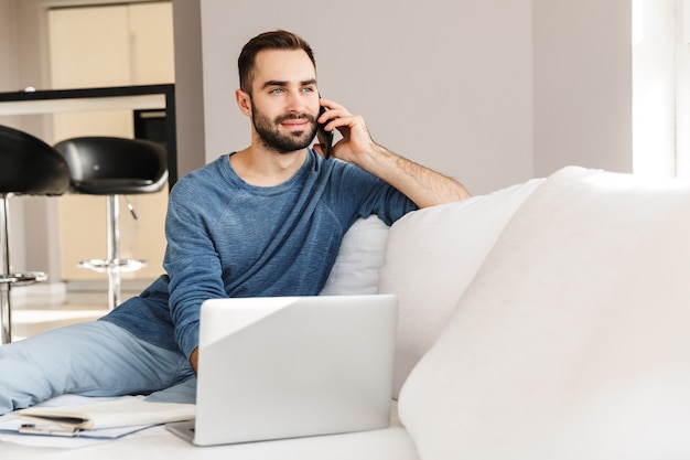 Jeune homme séduisant se reposant sur un canapé à la maison, travaillant sur un ordinateur portable, parlant sur un téléphone portable