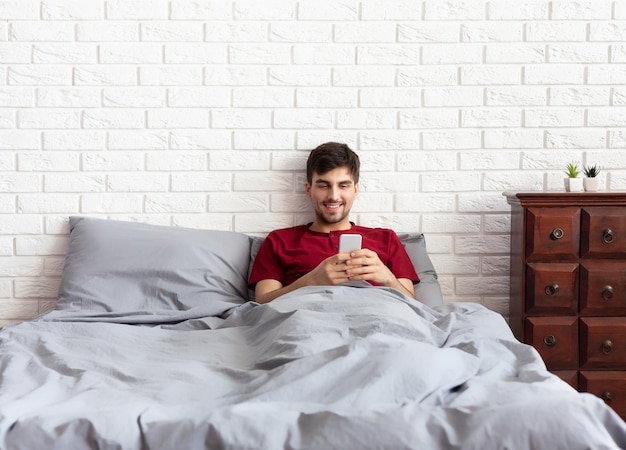 Jeune homme séduisant utilisant un smartphone et envoyant des SMS en position couchée dans son lit