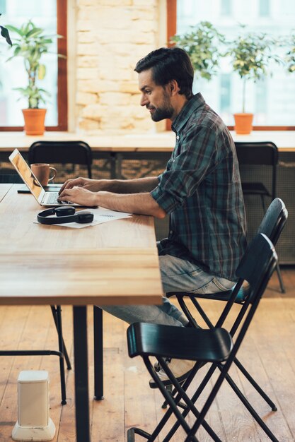 Jeune homme séduisant en vêtements décontractés utilisant un ordinateur portable moderne assis seul