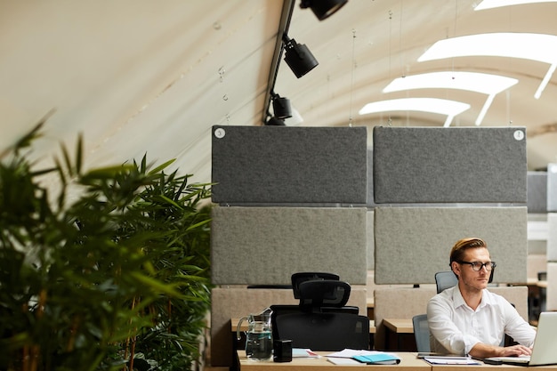 Jeune homme sérieux concentré sur un projet d'entreprise assis au bureau et utilisant un ordinateur portable dans un bureau moderne