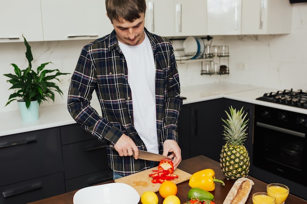 Jeune homme sérieux debout près de la table tenant un couteau et coupe le poivron rouge.