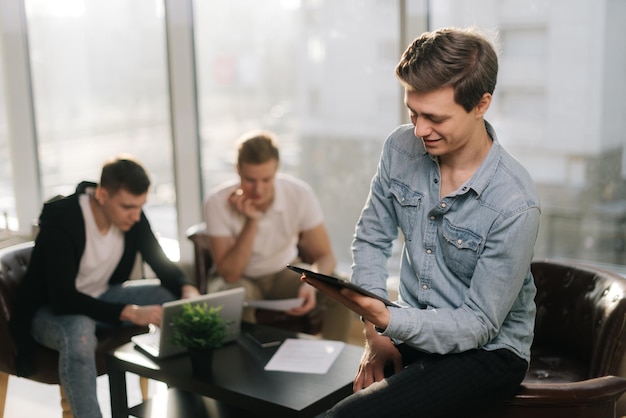 Un jeune homme sérieux en démarrage d'entreprise utilise une tablette sur fond de collègues de travail et de lumière du soleil Concept de travail d'équipe de jeunes gens d'affaires