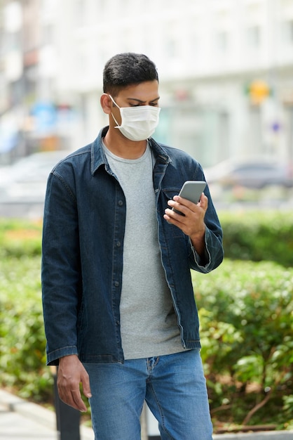 Jeune homme sérieux portant un masque médical marchant dans la rue et lisant des nouvelles sur le coronavirus sur smartphone