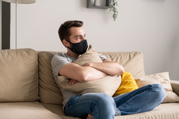 Jeune homme solitaire en masque en tissu assis avec un oreiller sur un canapé et pensant à la quarantaine des coronavirus