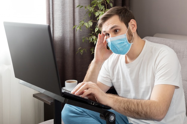 Jeune homme sombre caucasien en stress travaillant à domicile avec un masque de protection à l'aide d'un ordinateur portable et d'Internet. Coronavirus covid 19 quarantine. Travail à distance, pigiste, bureau à domicile sur canapé