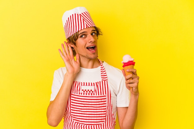 Jeune homme de sorbetière avec maquillage tenant une crème glacée isolée sur fond jaune essayant d'écouter un potin.