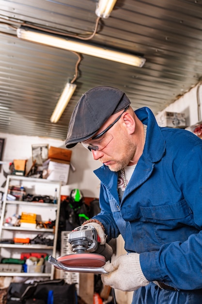 Un jeune homme soudeur dans un T-shirt bleu, des lunettes et des gants de construction processus métal une meuleuse d'angle dans le garage