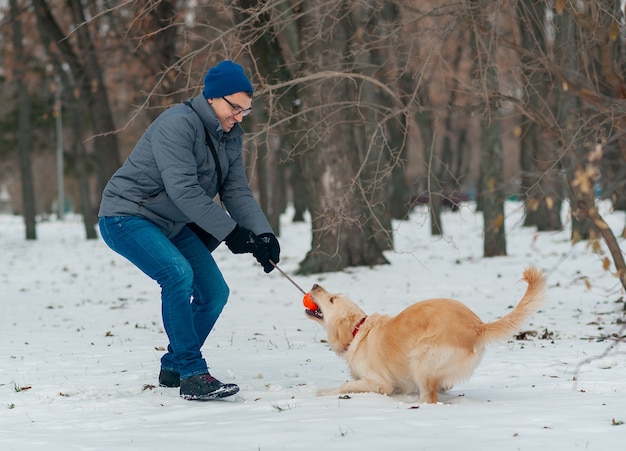 Jeune Homme Soufflant Des Flocons De Neige De Ses Mains à Son Chien Golden Retriever Dans Une Journée D'hiver. Amitié, Animal De Compagnie Et Humain.