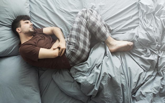 Jeune homme souffrant de maux d'estomac, allongé dans son lit à la maison, vue de dessus. Notion de problèmes de santé