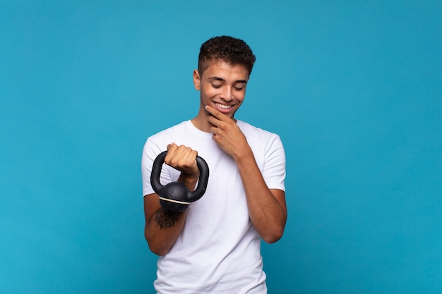 Jeune homme soulevant un haltère souriant avec une expression heureuse et confiante avec la main sur le menton, se demandant et regardant sur le côté