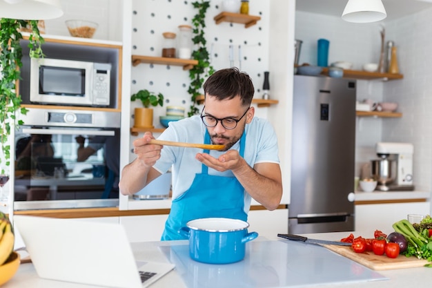 Jeune homme souriant appréciant le goût et l'odeur tout en cuisinant le déjeuner dans la cuisine