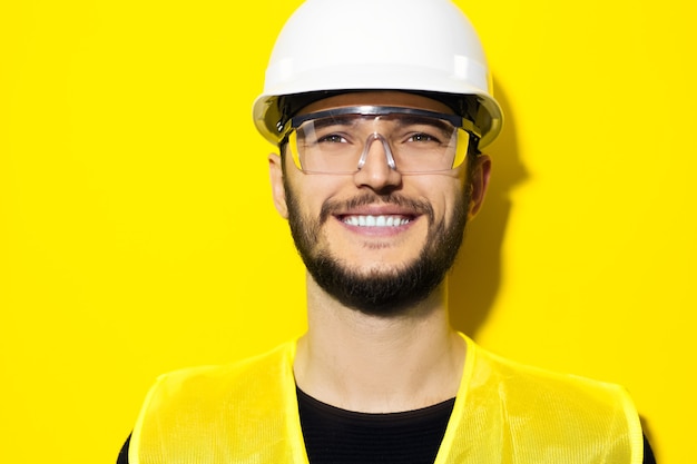 Jeune homme souriant architecte, ingénieur constructeur, portant des lunettes de sécurité de construction, casque dur et veste réfléchissante sur mur jaune