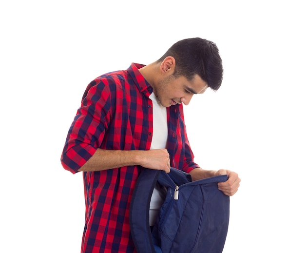 Jeune homme souriant aux cheveux noirs en T-shirt blanc et chemise à carreaux rouge tenant un sac à dos bleu