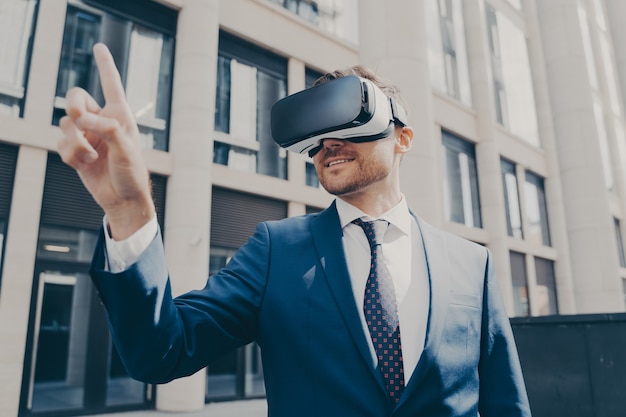 Jeune homme souriant et beau entrepreneur avec du chaume, vêtu d'une tenue de soirée, portant des lunettes VR portables, essayant de toucher quelque chose en réalité virtuelle avec son doigt, seul dans la rue de la ville