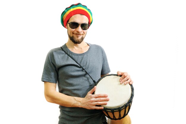 Jeune homme souriant en chapeau rasta et t-shirt gris sur fond blanc avec tambour africain djembé