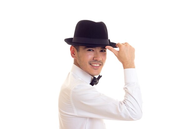 Jeune homme souriant en chemise blanche avec noeud papillon noir et chapeau noir sur fond blanc en studio