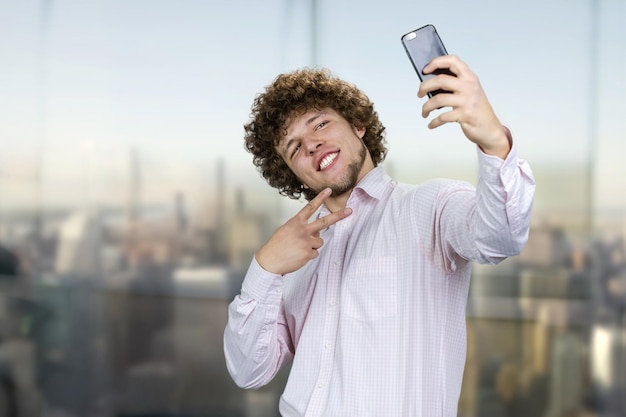 Photo jeune homme souriant avec les cheveux bouclés faisant un selfie avec le geste de victoire signe paysage urbain dans le