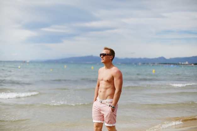 Un Jeune Homme Souriant Avec Un Corps Athlétique Pose Sur La Plage Près De L'océan En Journée D'été. Concept De Vacances. Mode De Vie Sportif.