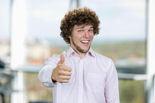 Photo un jeune homme souriant heureux aux cheveux bouclés montre le pouce vers le haut de la fenêtre intérieure en arrière-plan