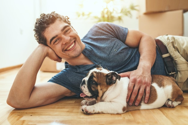 Un jeune homme souriant et heureux est allongé sur un sol dans le salon de son nouvel appartement et tient un petit chiot.