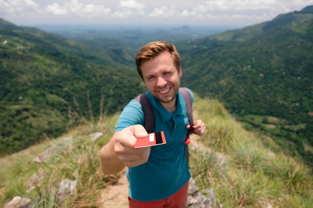 Jeune homme souriant montrant une carte de crédit vide, il se tient au sommet d'une montagne et montre l'outdo...