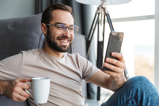 un jeune homme souriant et optimiste à l'intérieur à la maison en train de boire du café à l'aide d'un téléphone portable.