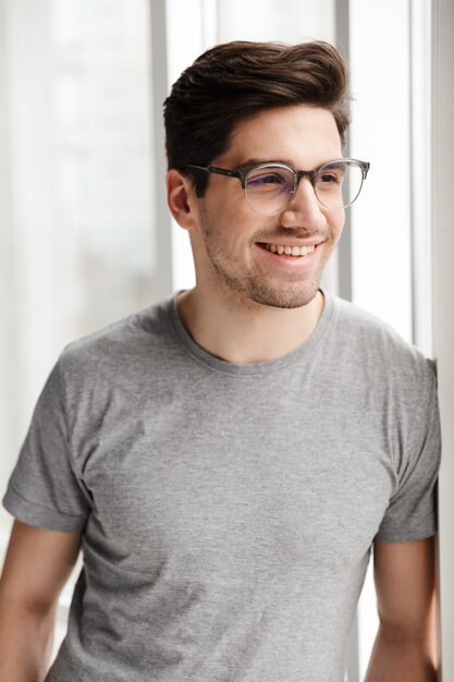 un jeune homme souriant et positif à l'intérieur à la maison en regardant la fenêtre.