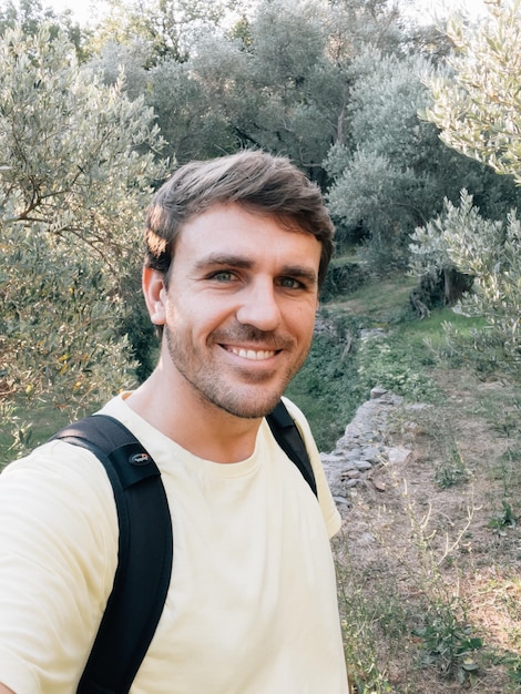 Un jeune homme souriant avec un sac à dos se tient dans une oliveraie