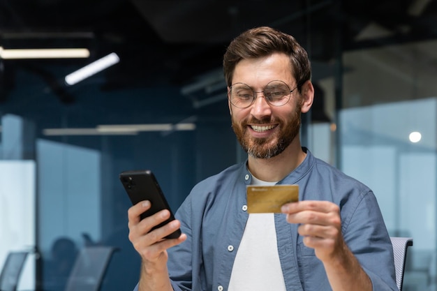 Un jeune homme souriant tient un téléphone et une carte de crédit se trouve dans le bureau compose au téléphone