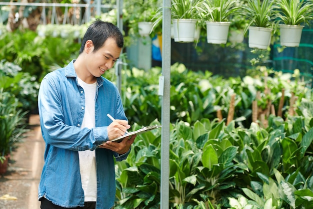 Jeune homme souriant travaillant dans une pépinière, comptant les plantes et prenant des notes dans un document