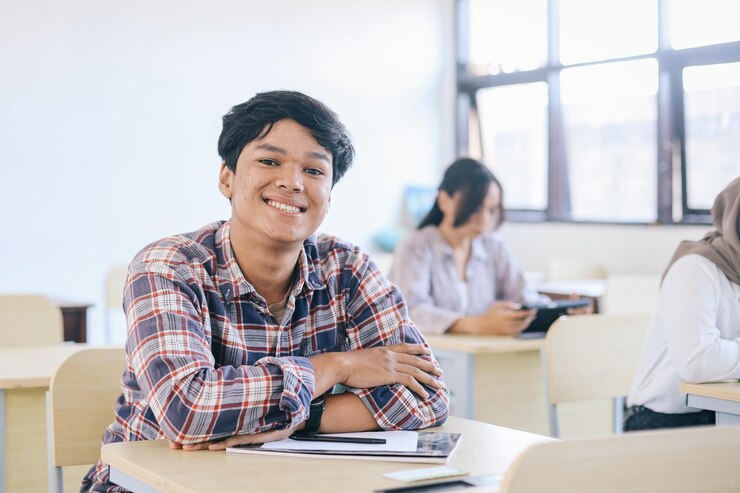 Un jeune homme souriant à l'université s'appuie sur un bureau avec des camarades de classe en arrière-plan