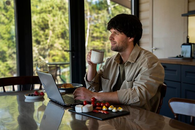 Un jeune homme souriant utilise un ordinateur portable et prend son petit déjeuner dans la cuisine.