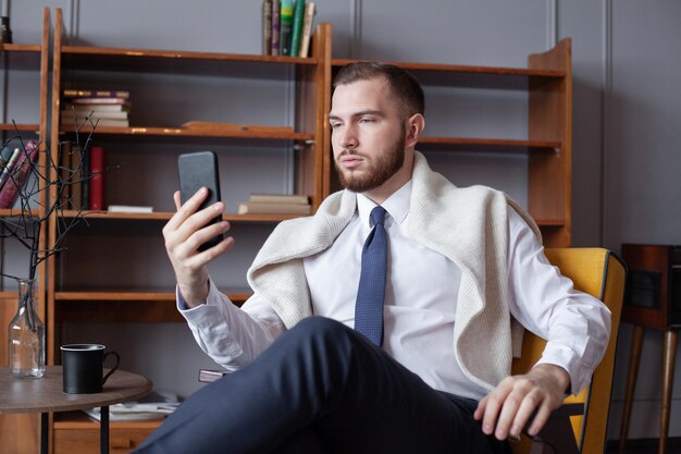 Un jeune homme souriant en vêtements décontractés à la mode se fait un selfie sur un téléphone portable.