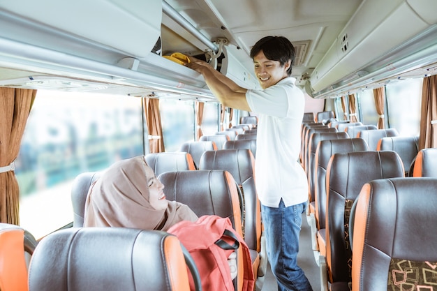 Un jeune homme sourit alors qu'il aide une femme portant un foulard à mettre son sac sur une étagère en se tenant debout dans le bus