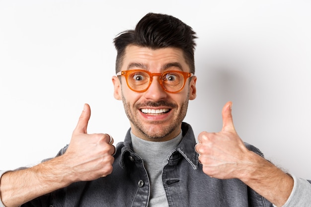 Jeune homme de soutien dans des verres souriant, montrant les pouces vers le haut pour louer ou approuver une bonne chose, debout sur un fond blanc.