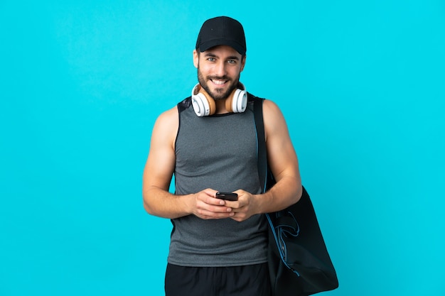 Jeune homme de sport avec sac de sport isolé sur mur bleu envoyant un message avec le mobile