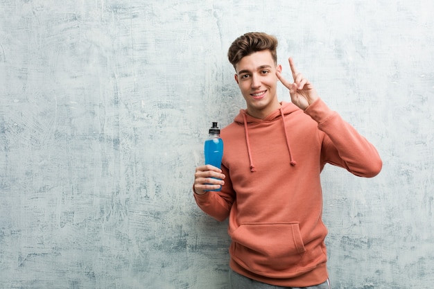 Jeune homme de sport tenant une boisson énergisante montrant le signe de la victoire et souriant largement.