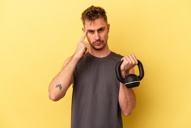 Jeune homme de sport tenant un haltère isolé sur fond jaune pointant le temple avec le doigt, pensant, concentré sur une tâche.