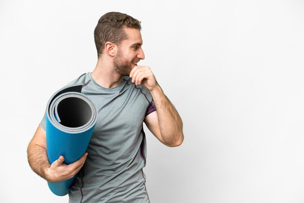 Jeune homme sportif allant à des cours de yoga tout en tenant un tapis sur fond blanc isolé en pensant à une idée et en regardant de côté