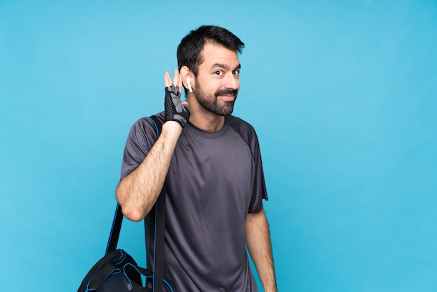 Jeune homme sportif avec barbe sur bleu isolé, écoutant quelque chose en mettant la main sur l'oreille