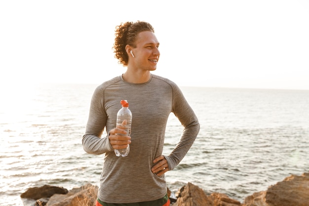 Jeune homme sportif debout sur la plage de l'eau potable en écoutant de la musique.