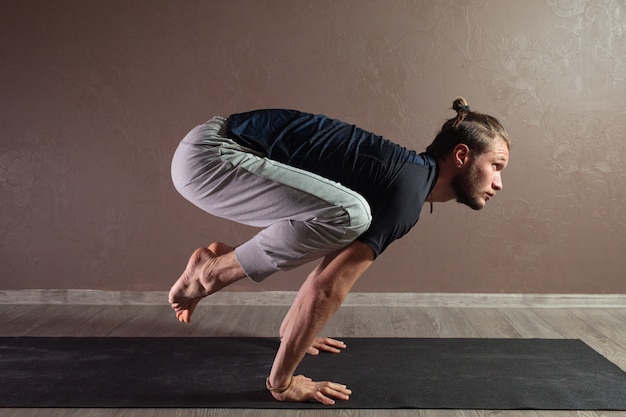 Jeune homme sportif pratiquant le yoga, méditant dans une pose de demi-lotus, travaillant, portant des vêtements de sport, intérieur pleine longueur, studio de yoga brun