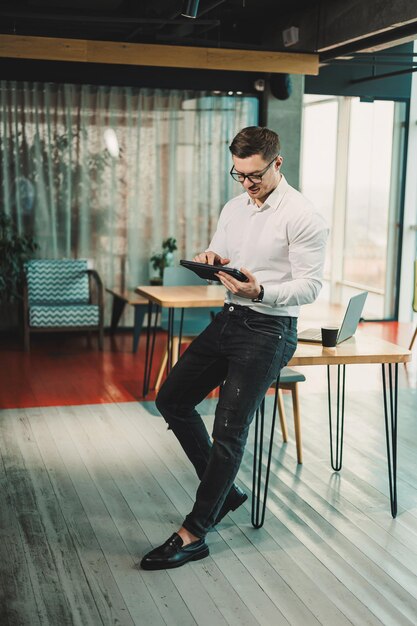 Un jeune homme à succès en lunettes et en chemise blanche travaille sur une tablette sur un projet dans un espace de travail moderne.