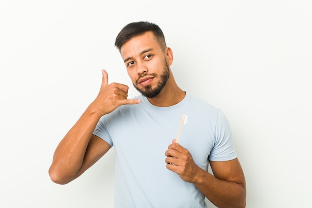 Jeune homme sud-asiatique tenant une brosse à dents montrant un geste d'appel de téléphone mobile avec les doigts.